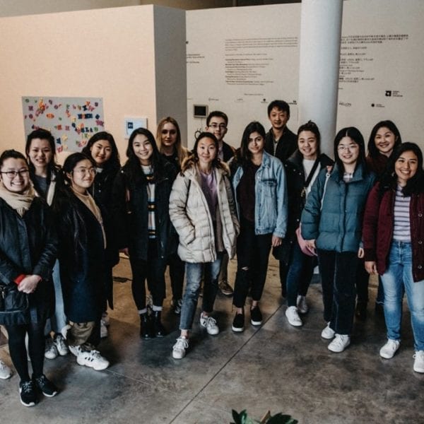 16 students pose in front of the exhibit wall