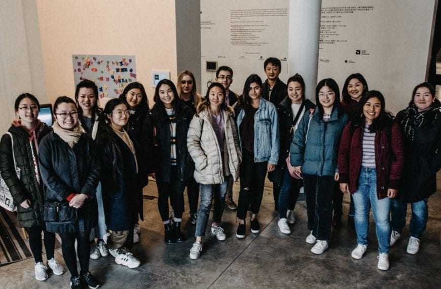 16 students pose in front of the exhibit wall