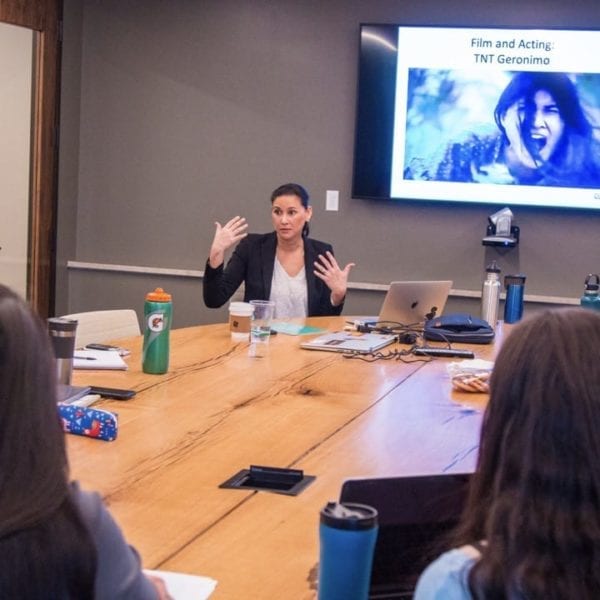 Tailinh Agoyo speaks to the class, seated around a seminar table, with an image from TNT's Geronimo on a screen behind her.