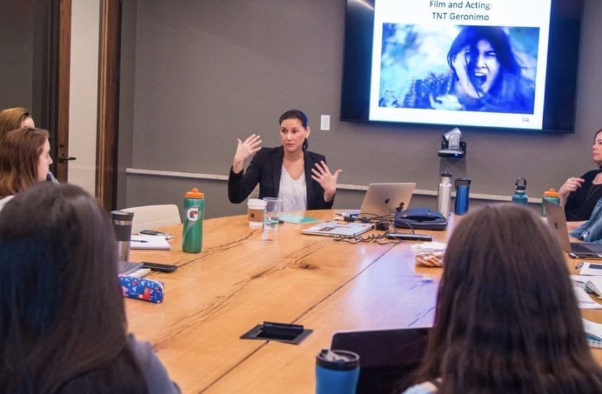 Tailinh Agoyo speaks to the class, seated around a seminar table, with an image from TNT's Geronimo on a screen behind her.