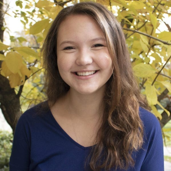 A headshot of Sarah Jesup wearing a blue v-neck t-shirt standing in front of a tree with yellow fall leaves