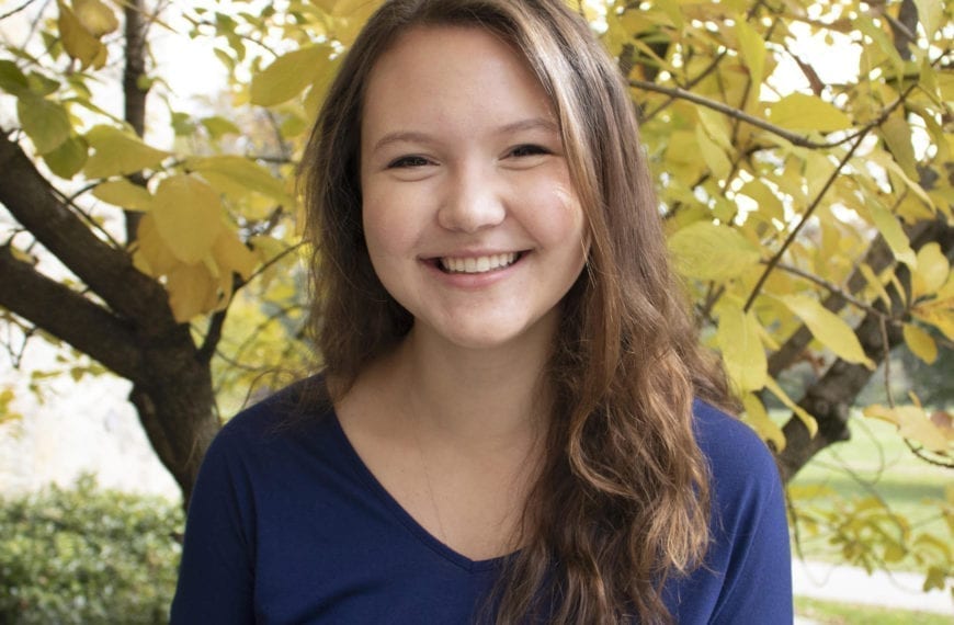 A headshot of Sarah Jesup wearing a blue v-neck t-shirt standing in front of a tree with yellow fall leaves