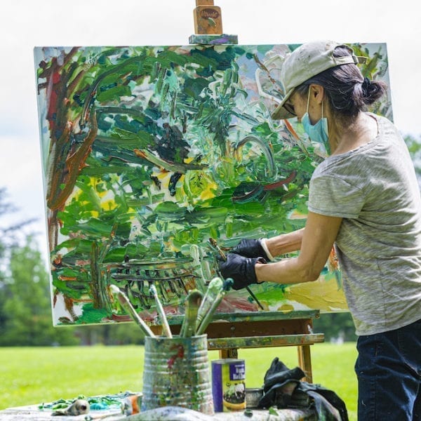 Ying Li, wearing a mask and hat, paints outside by the Duck Pond.
