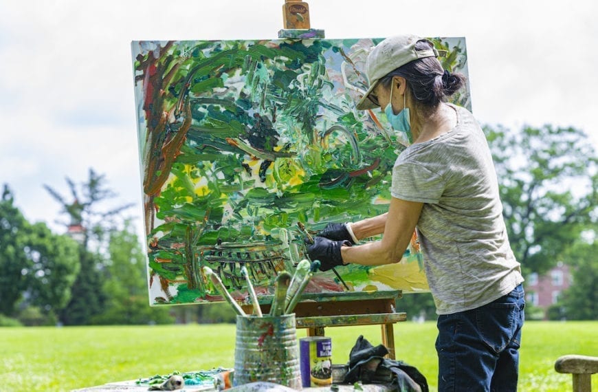 Ying Li, wearing a mask and hat, paints outside by the Duck Pond.