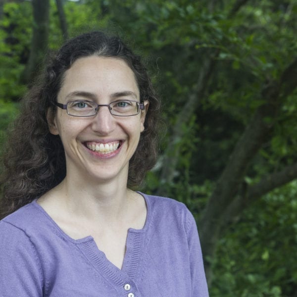 Sarah Horowitz, wearing a purple shirt and glasses, stands in front of a green tree on the Haverford campus