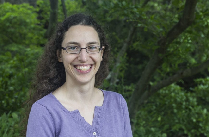Sarah Horowitz, wearing a purple shirt and glasses, stands in front of a green tree on the Haverford campus