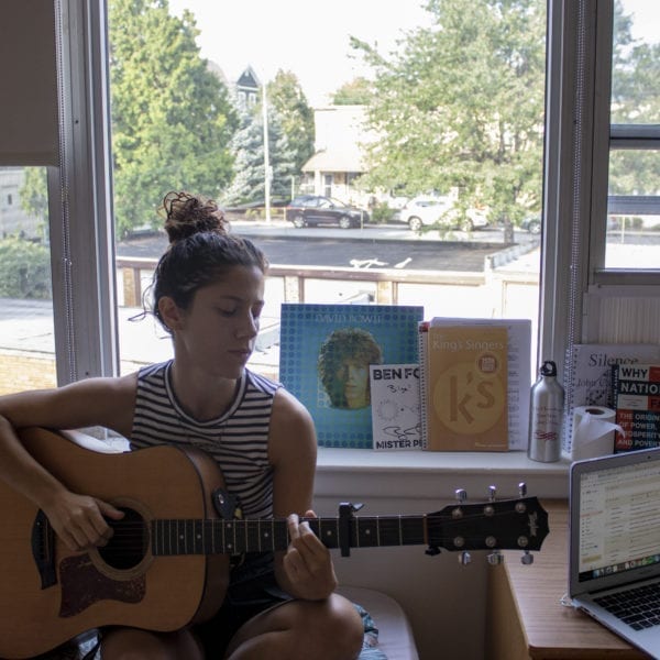 Bilge Nur Yilmaz plays acoustic guitar sitting on her bed next to her open laptop and in front of a David Bowie record