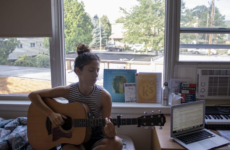 Bilge Nur Yilmaz plays acoustic guitar sitting on her bed next to her open laptop and in front of a David Bowie record