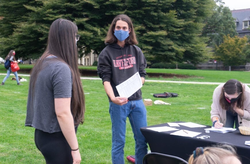 A student wearing a mask and a Haverford sweatshirt hands another student voter-registration paperwork at an event on Founders Green.