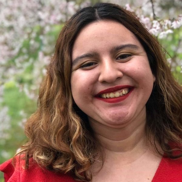 Person in red shirt with shoulder length hair smiles in front of flowering tree