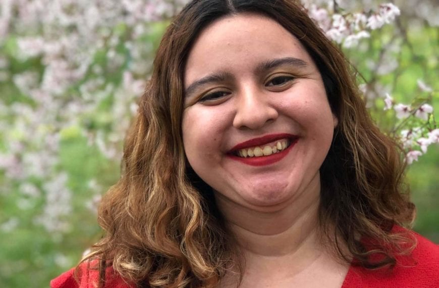 Person in red shirt with shoulder length hair smiles in front of flowering tree