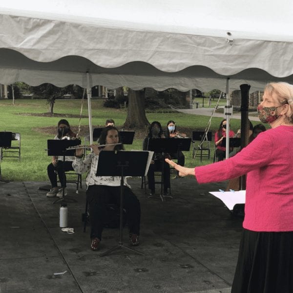 woman with a face mask directs an ensemble of people playing instruments outside under a tent