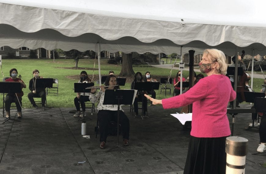 woman with a face mask directs an ensemble of people playing instruments outside under a tent