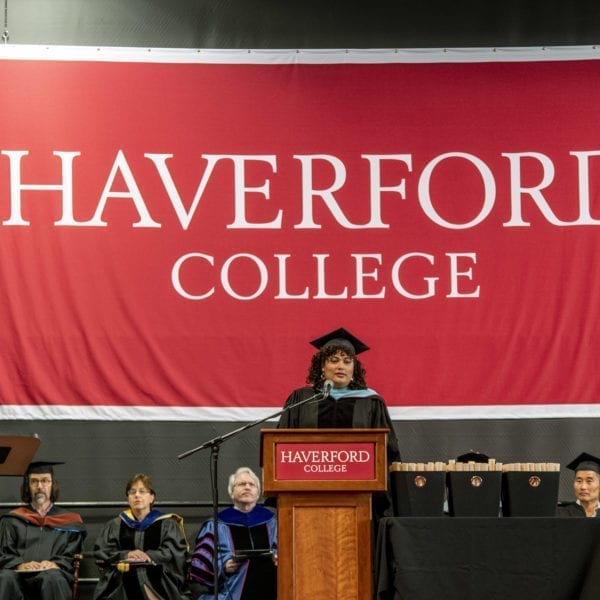 Raquel Esteves-Joyce speaking at a podium in front of a large, red Haverford College sign on the stage at Commencement