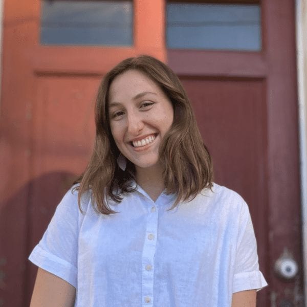Woman in a white button up, short-sleeved shirt smiling in front of a dark red door.