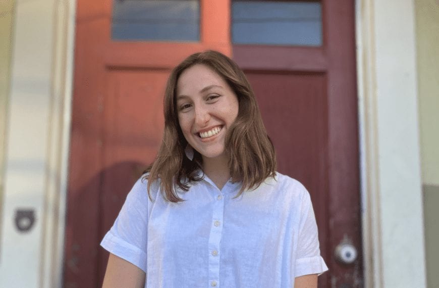 Woman in a white button up, short-sleeved shirt smiling in front of a dark red door.