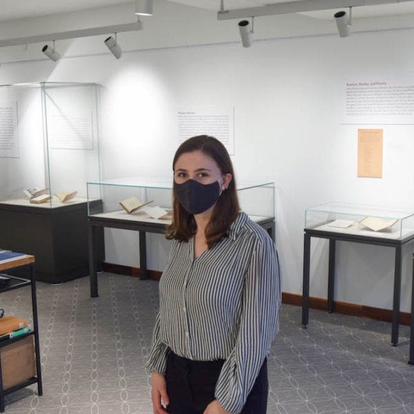Charlotte Scott, wearing a mask, stands amid display cases in the exhibit she curated in the library