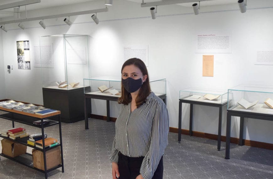 Charlotte Scott, wearing a mask, stands amid display cases in the exhibit she curated in the library