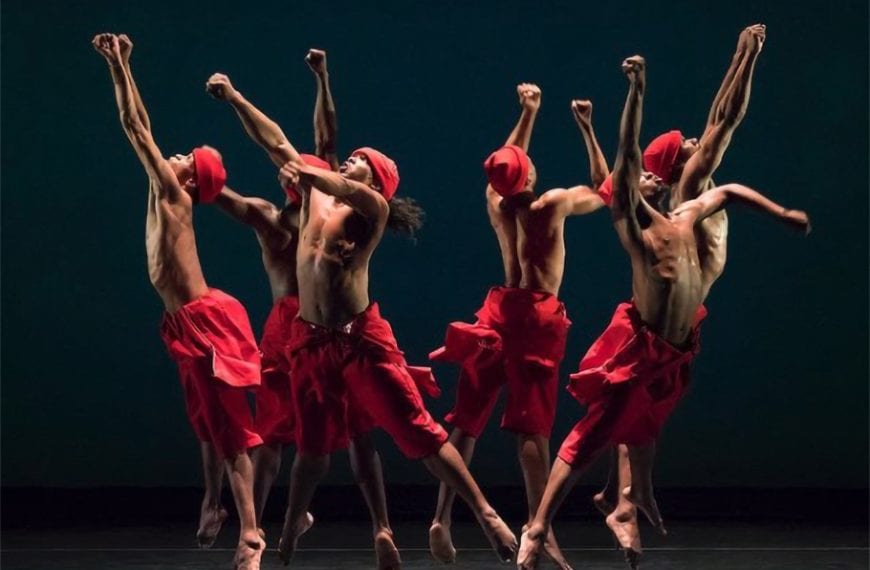 Six men in red headwraps and red wrap pants leap in a circle with one fist up