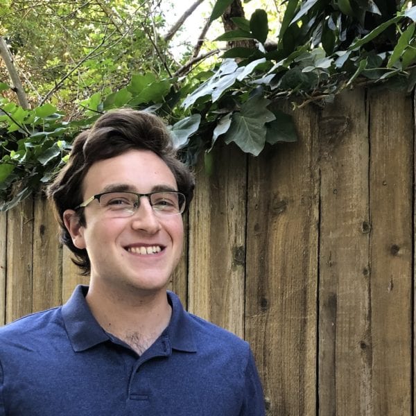 Colin smiles in a blue shirt in front of a wooden fence.