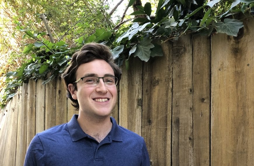 Colin smiles in a blue shirt in front of a wooden fence.