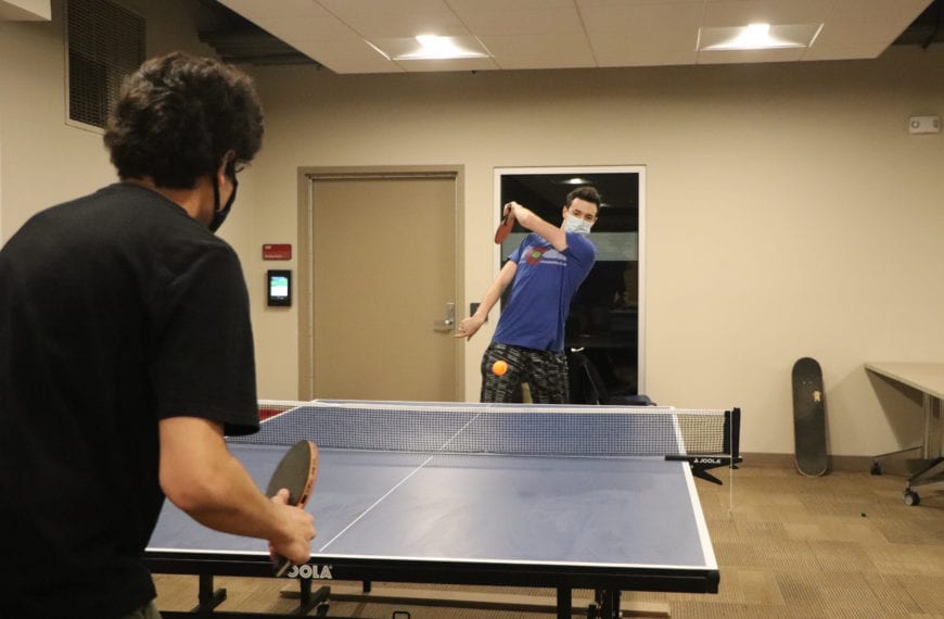 A student his a ping pong ball to another student, who crouches ready to hit it back.