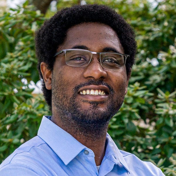 Professor Clyde Daly, wearing a blue shirt, stands in front of a background of leaves and trees.