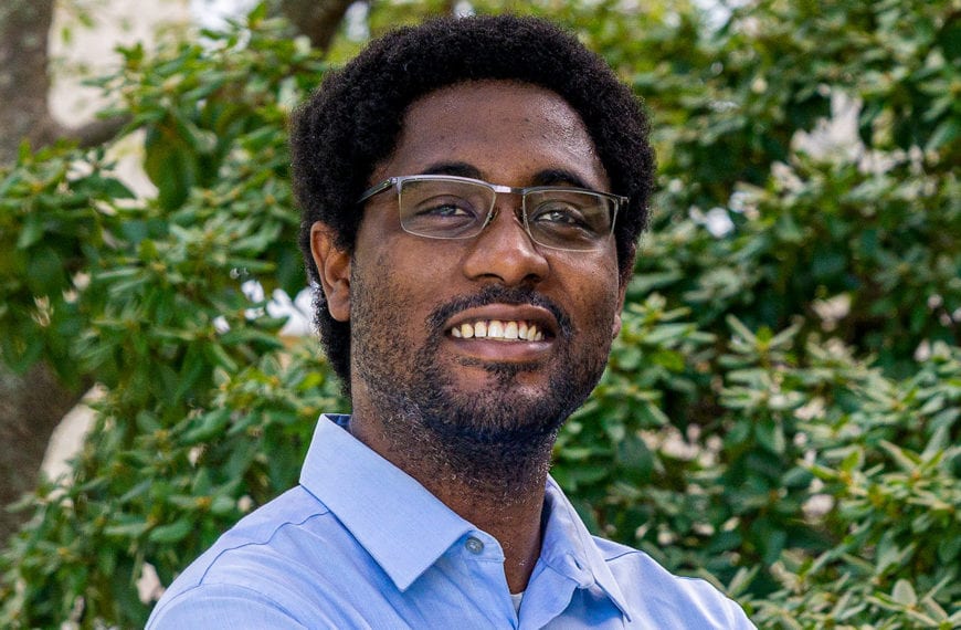 Professor Clyde Daly, wearing a blue shirt, stands in front of a background of leaves and trees.