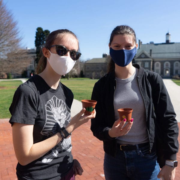 Two students in masks stand on Founders Green holding plants