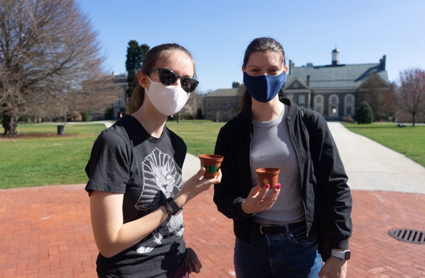 Two students in masks stand on Founders Green holding plants