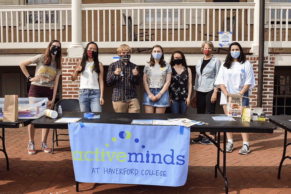 Students and President Wendy Raymond, all wearing masks, stand behind a table with an Active Minds banner on it in front of Founders Hall
