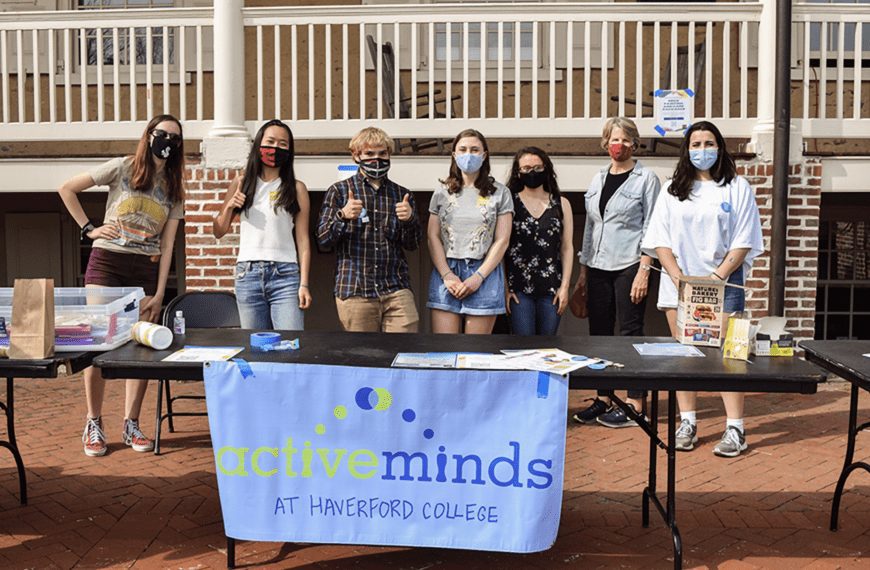 Students and President Wendy Raymond, all wearing masks, stand behind a table with an Active Minds banner on it in front of Founders Hall