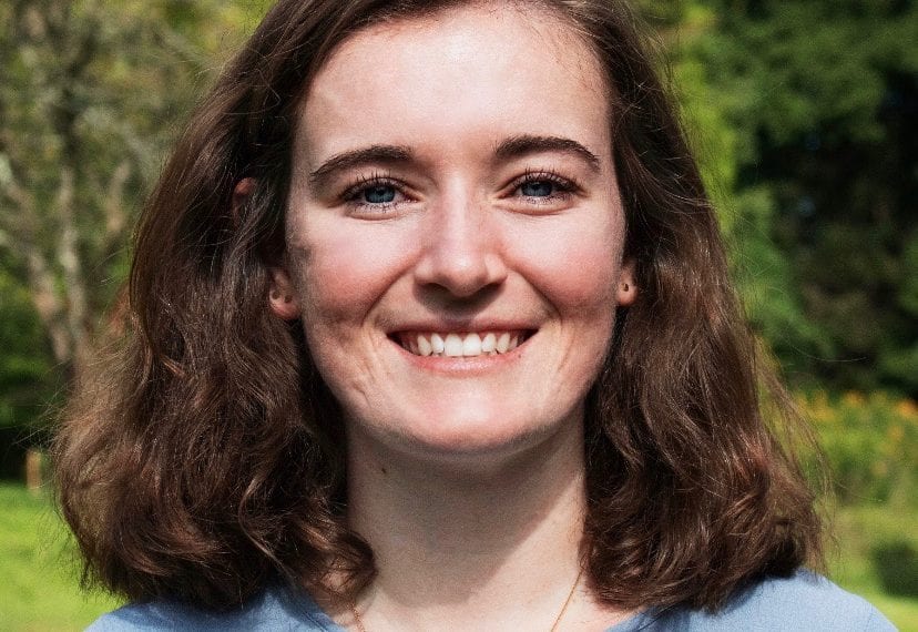 Annie Connolly-Sporing '20, wearing a blue shirt, stands in front of a background of trees.
