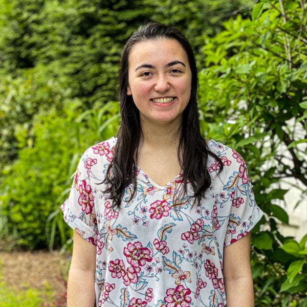 Elena smiles in front of a green bush, wearing a white and pink floral blouse.