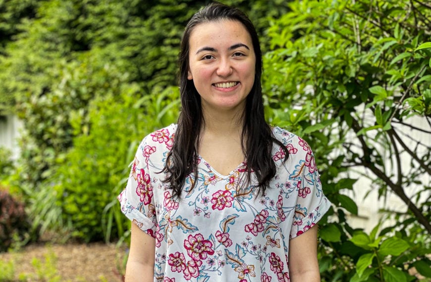 Elena smiles in front of a green bush, wearing a white and pink floral blouse.