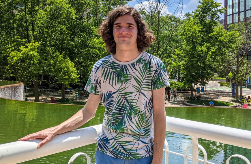 A headshot of Brett wearing a light colored shirt with palms on an overlook over a pond.