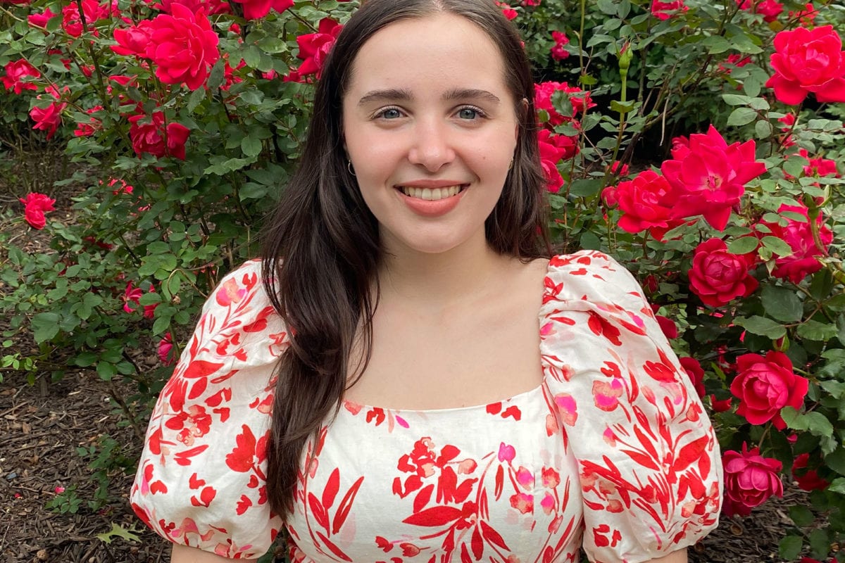 Sonia smiles in front of a rose bush wearing a white dress with a floral red design.