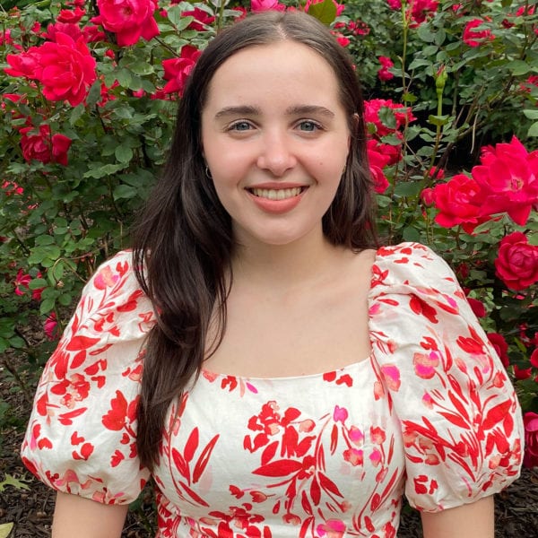 Sonia smiles in front of a rose bush wearing a white dress with a floral red design.