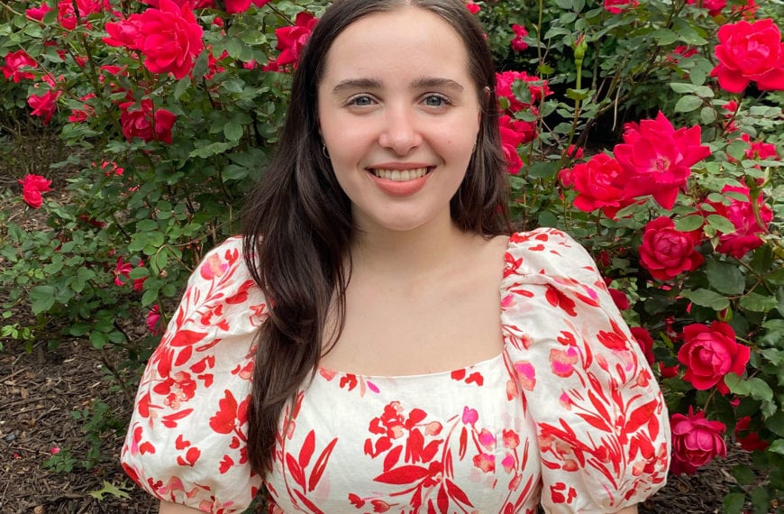 Sonia smiles in front of a rose bush wearing a white dress with a floral red design.