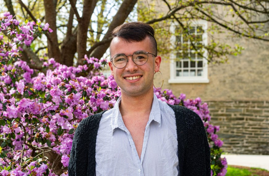 Headshot of Robbie wearing a striped shirt in front of a pink-purple bush on the side of Founders Hall.