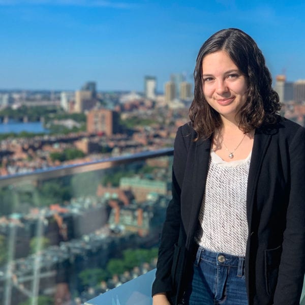 Headshot of Hannah in a patterned white shirt and suit jacket. In the background is a skyline.