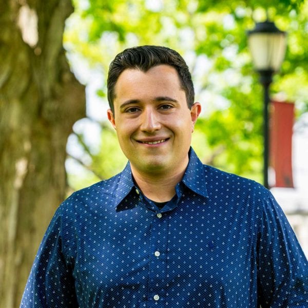 Headshot of Johnluca wearing a pattered blue shirt in front of a tree and the library.