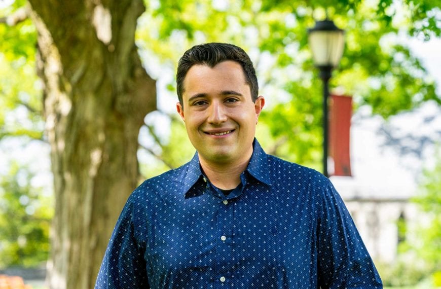 Headshot of Johnluca wearing a pattered blue shirt in front of a tree and the library.