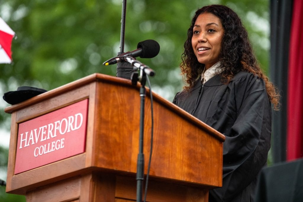 Praxedes Quintana wears a black robe and addresses the class from the podium.