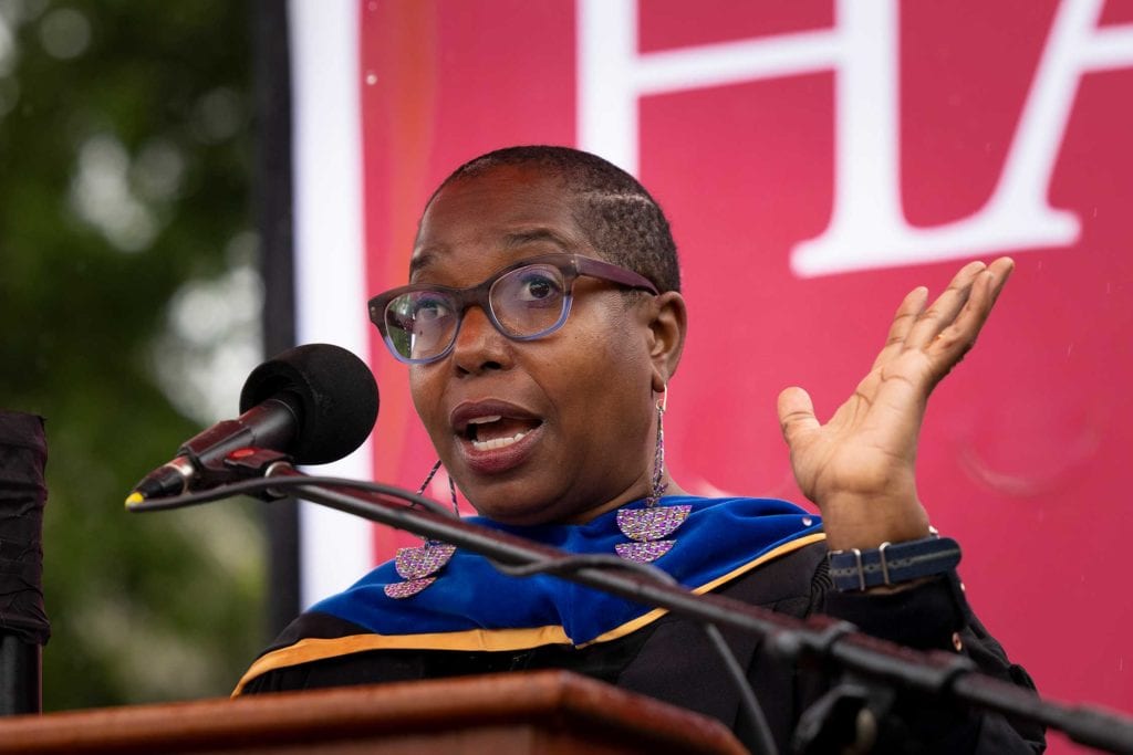 A close-up of Asali Solomon, wearing a black robe and blue hood and purple glasses, speaking at the podium.