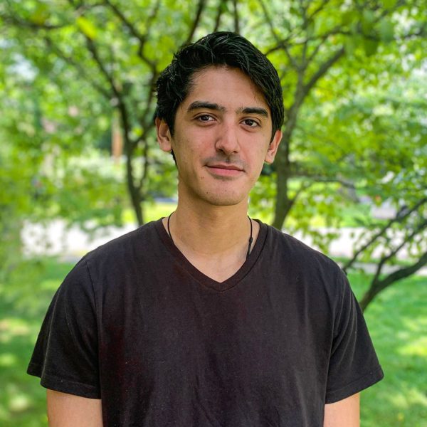 Benjamin Goldman wears a black t-shirt and stands under a tree