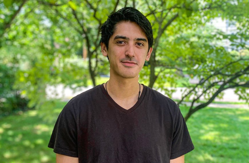 Benjamin Goldman wears a black t-shirt and stands under a tree
