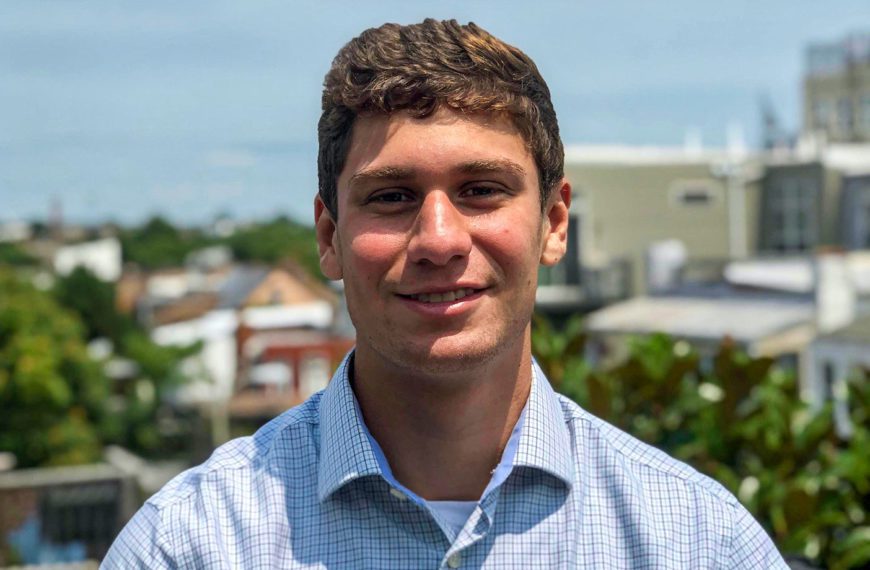 A picture of Brett wearing a blue button down with a city in the background.