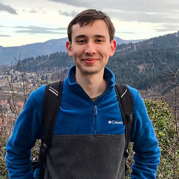 A photo of Colin in hiking clothes in front of a scenic valley.