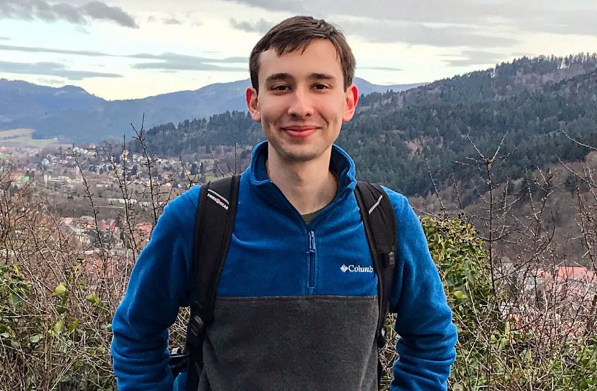 A photo of Colin in hiking clothes in front of a scenic valley.